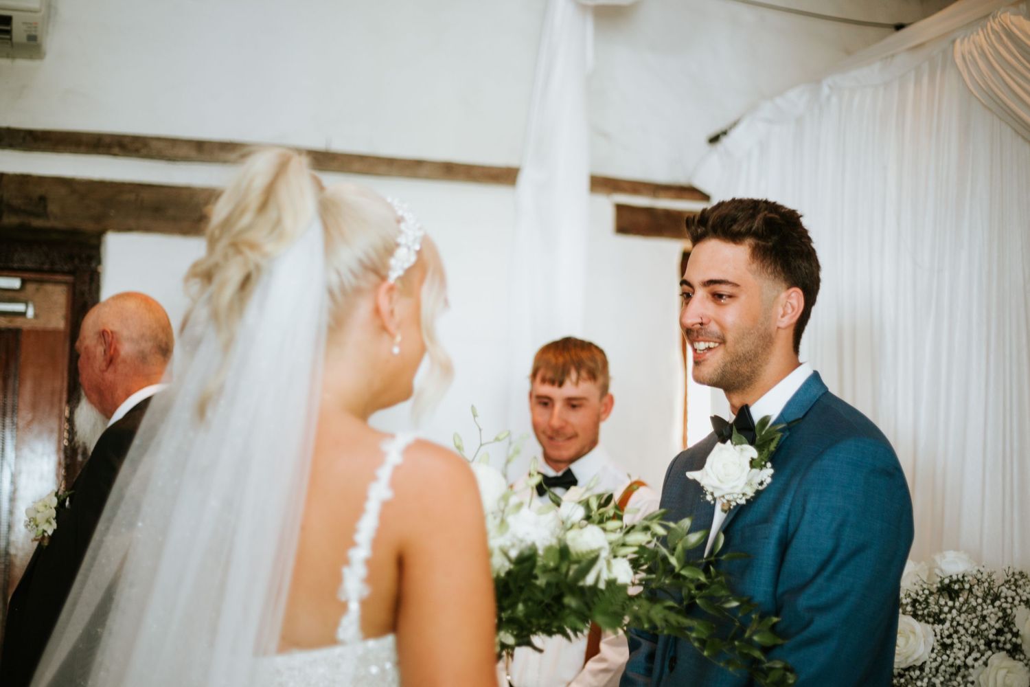 Groom in blue suit beaming at blonde bride holding greenery bouquet at wedding altar.