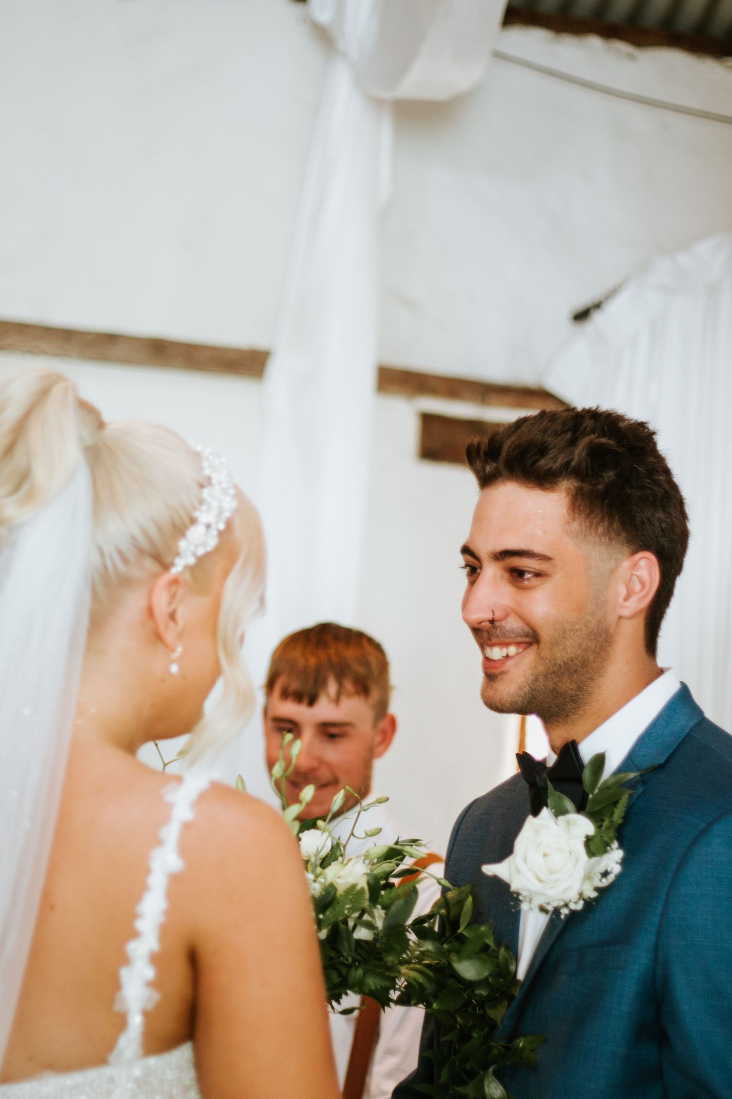 Groom in blue suit smiling at bride in white dress during indoor wedding ceremony.