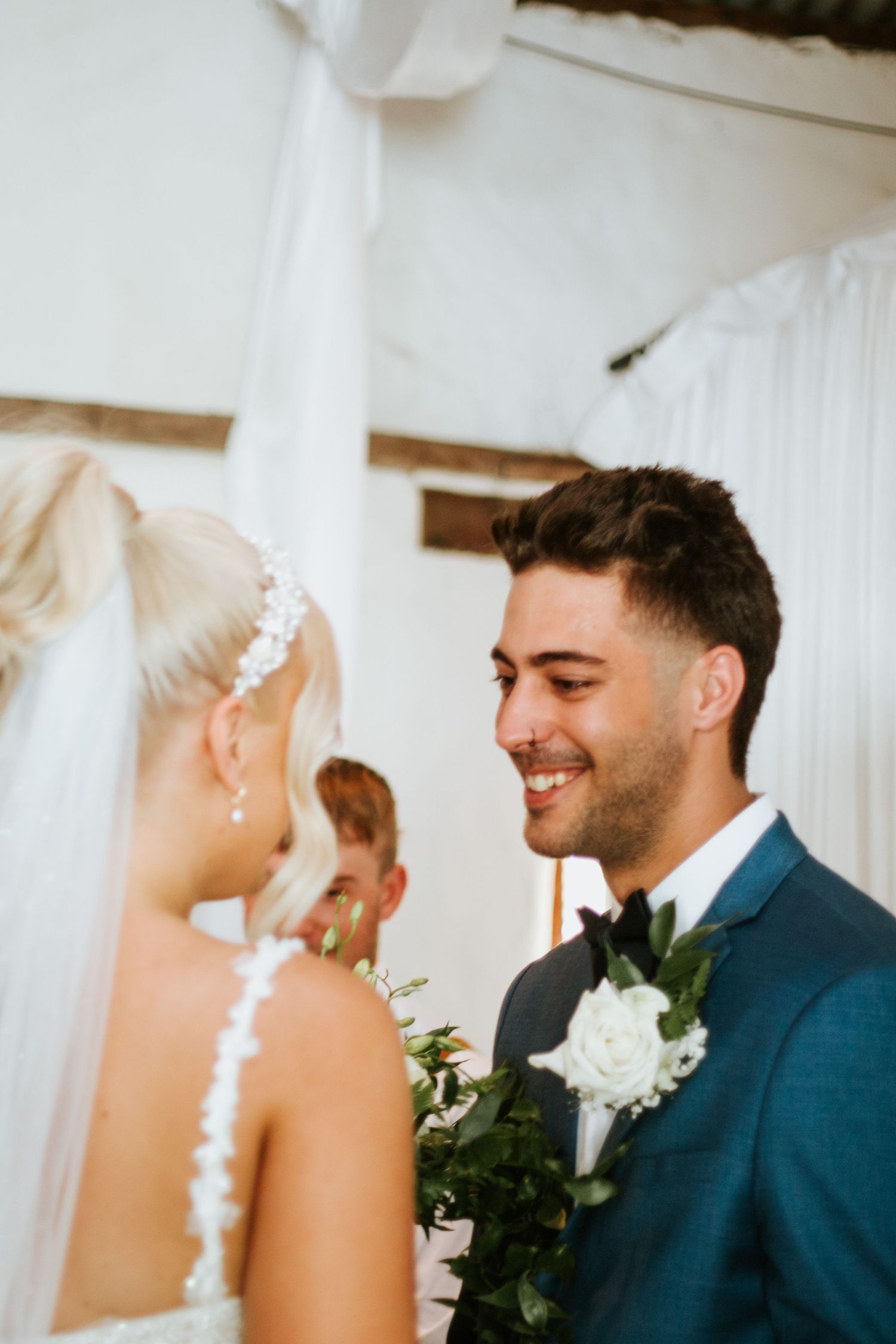 Smiling groom in blue suit gazes at blonde bride during intimate wedding ceremony.