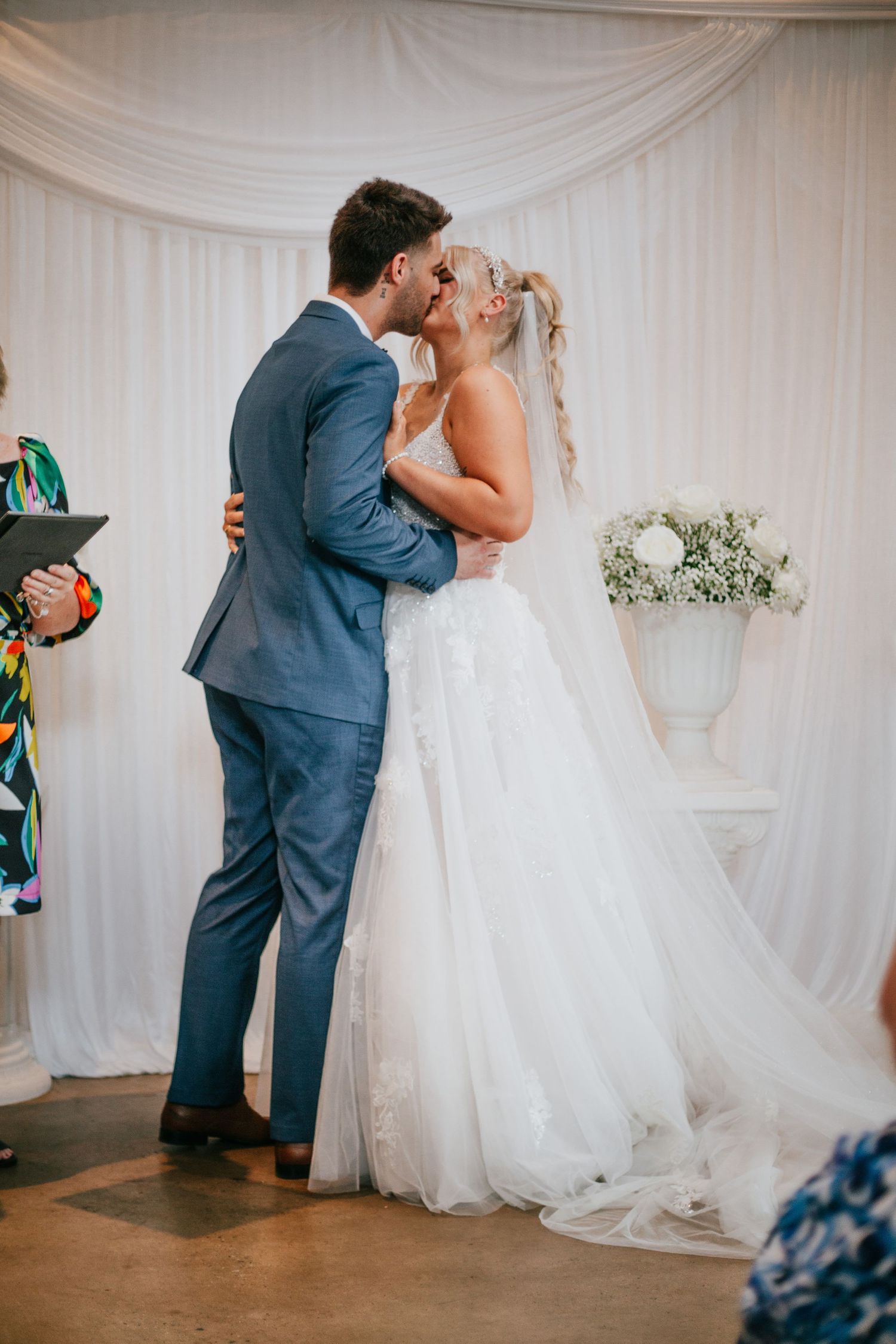 Bride and groom share first kiss at wedding ceremony in front of white drape backdrop.