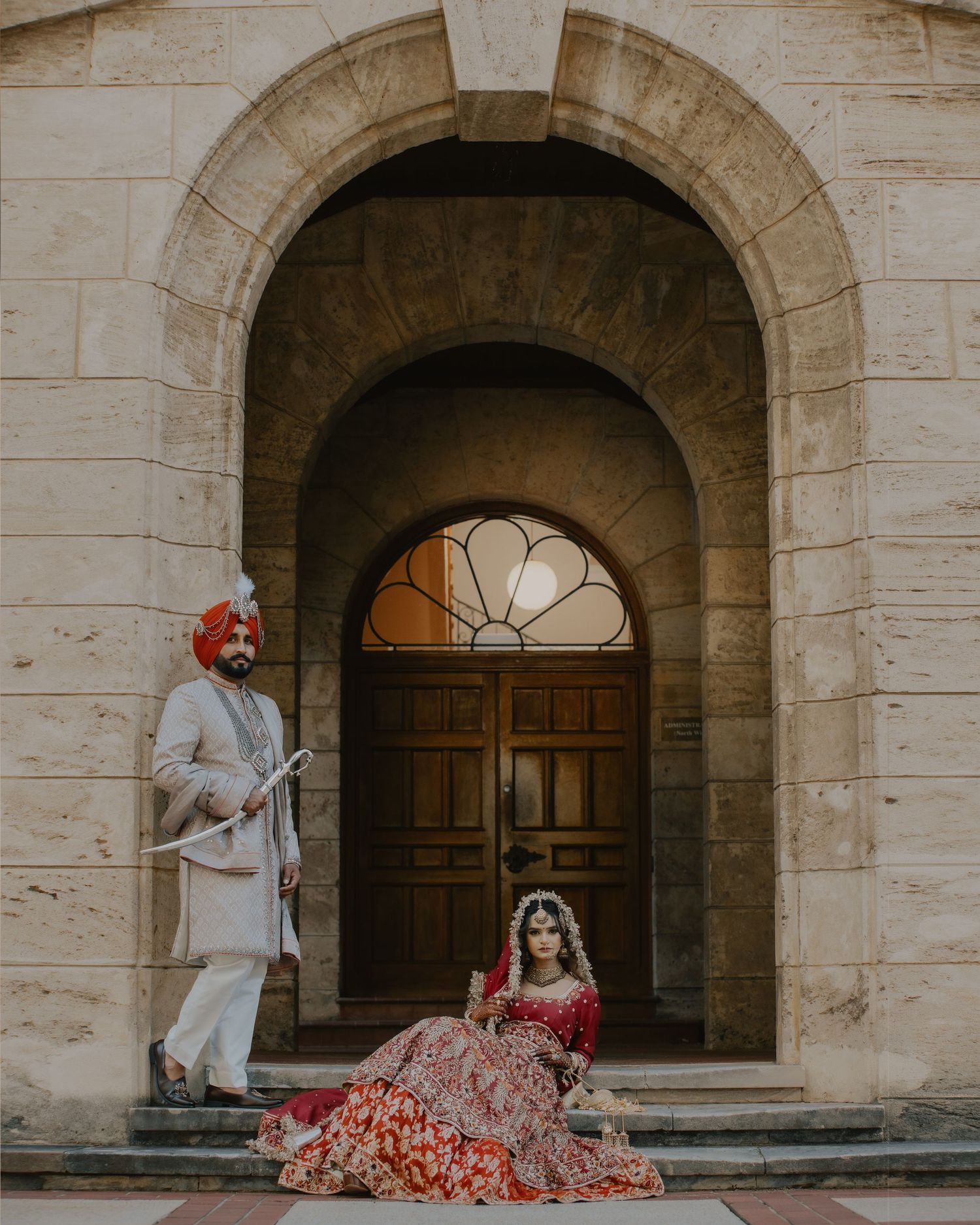A couple in traditional South Asian wedding attire poses beneath stone archways outside an ornate wooden door.