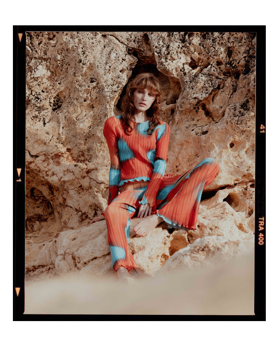Fashion model wearing a red and blue striped bodysuit poses against a rocky cave backdrop.