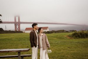 A couple stands on a grassy hill overlooking the Golden Gate Bridge in San Francisco on a foggy day.