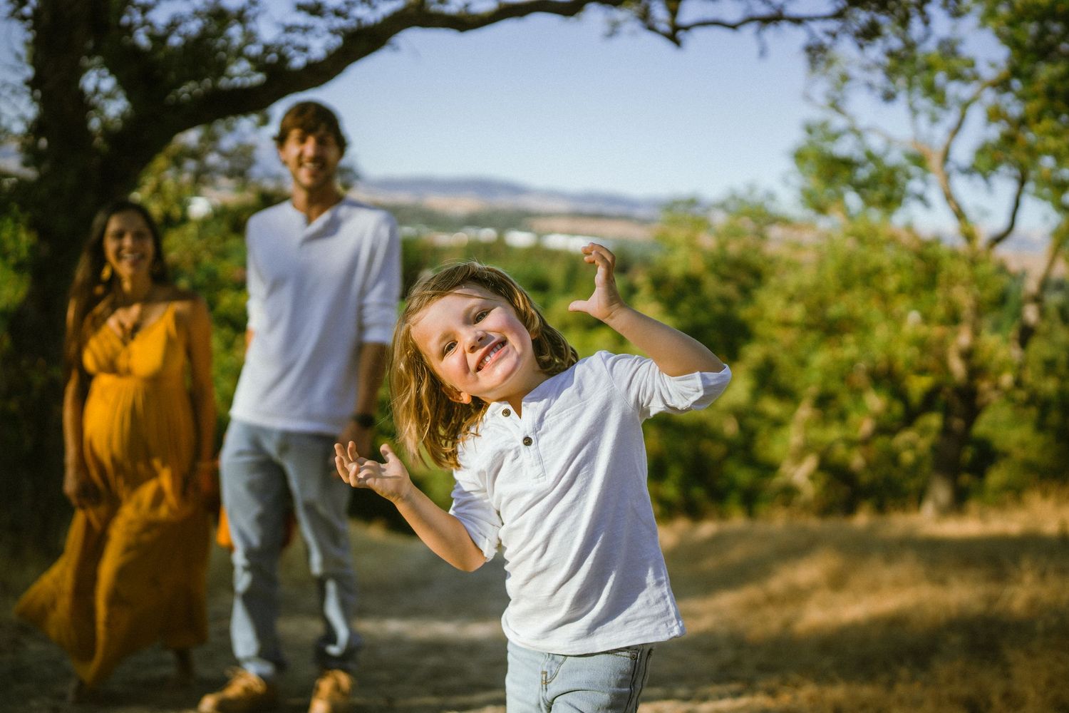 Joyful outdoor family photoshoot on a hillside with trees and mountains in the background.