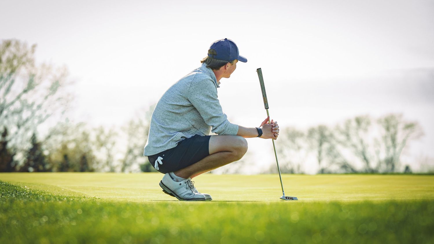 Golfer crouching on green grass to analyze a putt while holding putter on a sunny day.