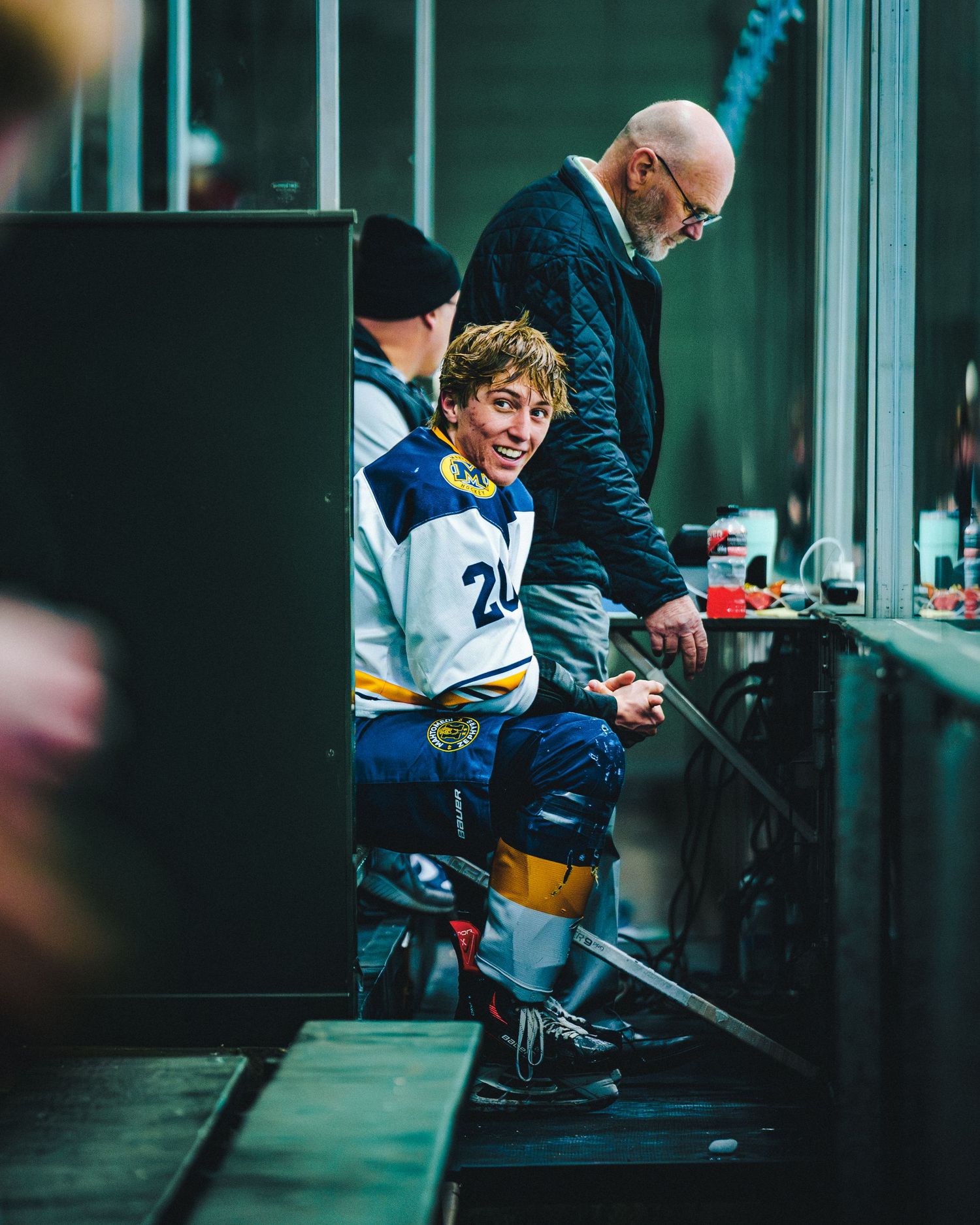 Hockey players and staff member interact at a player bench during a game with moody green lighting in the background.