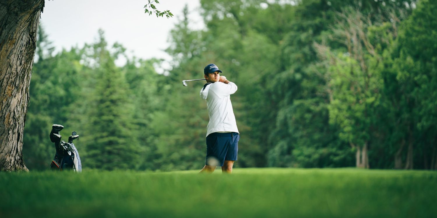 A golfer takes a swing on a lush green fairway with golf bag and trees in the background.