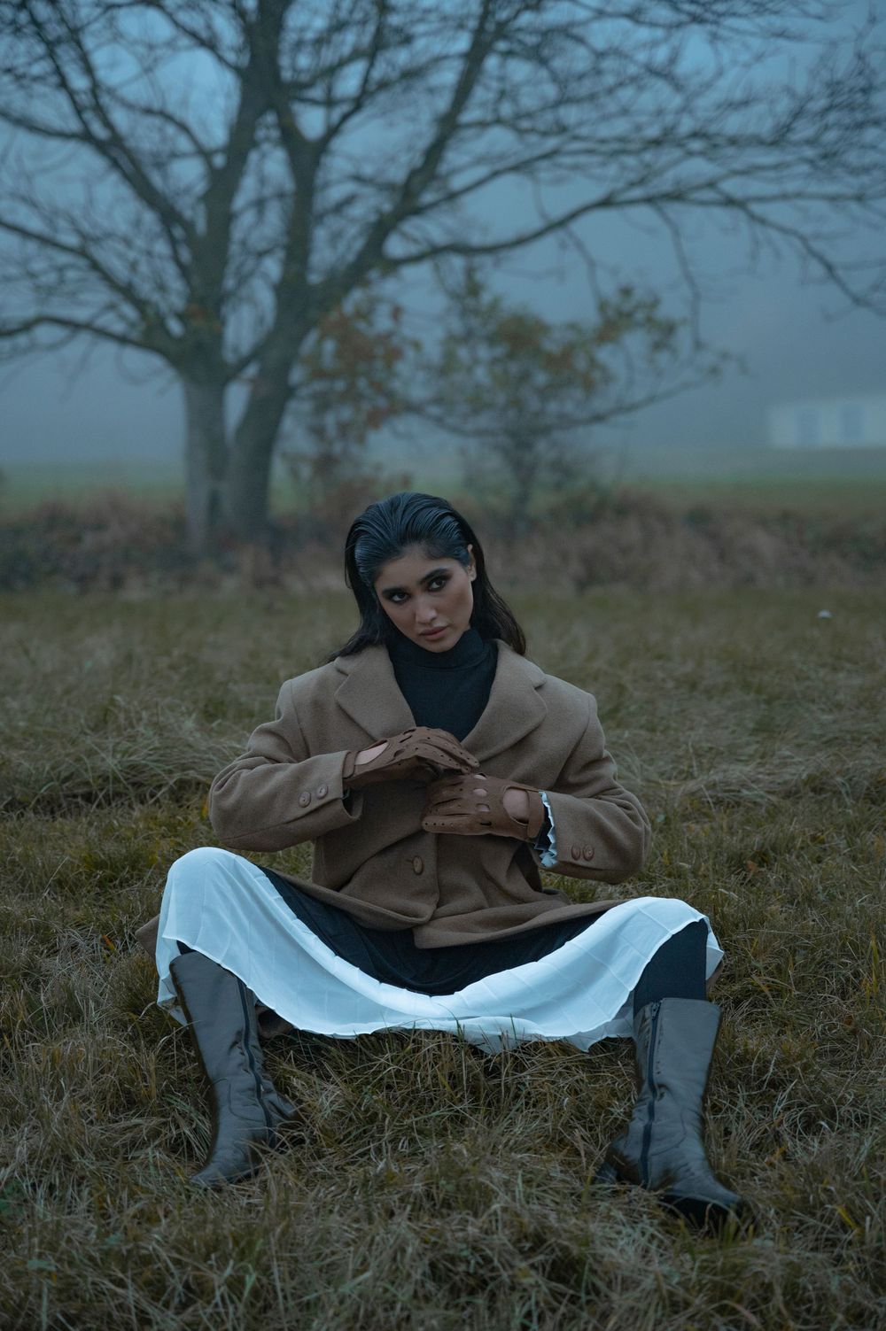 Woman in brown blazer and white skirt sits in foggy field wearing gray boots, looking at camera.
