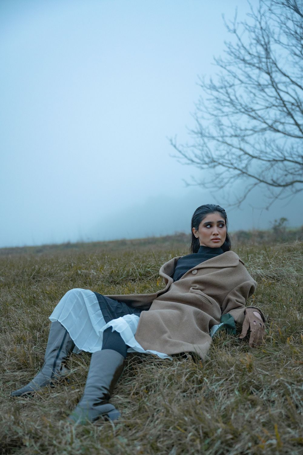 Woman in tan wrap coat and gray boots sitting in misty field with bare trees in background.