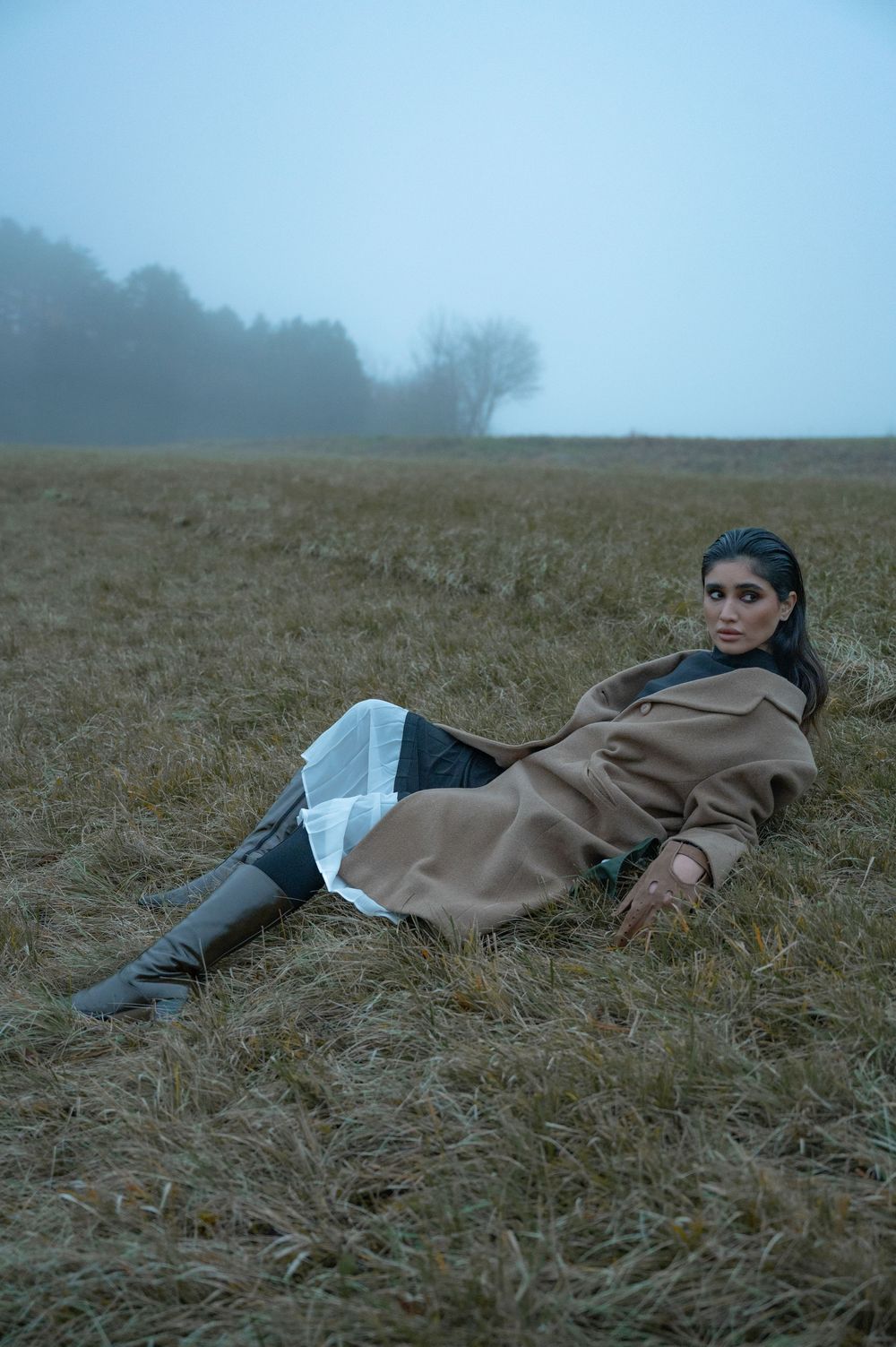 Woman in tan coat lying in misty field wearing dark boots, surrounded by dry grass with foggy trees in background.