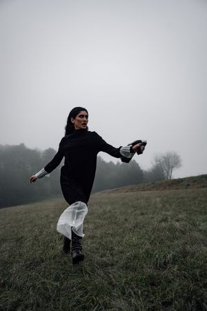 Woman in black dress running through misty foggy field, holding fabric, trees in background on overcast day.