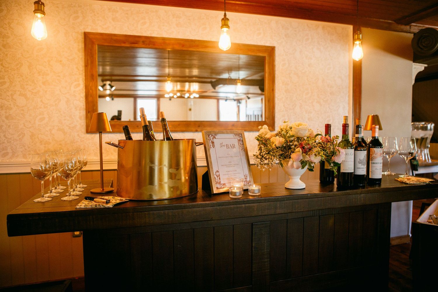 Elegant bar setup with wine bottles, champagne buckets, and glassware on a dark wood counter with warm lighting.