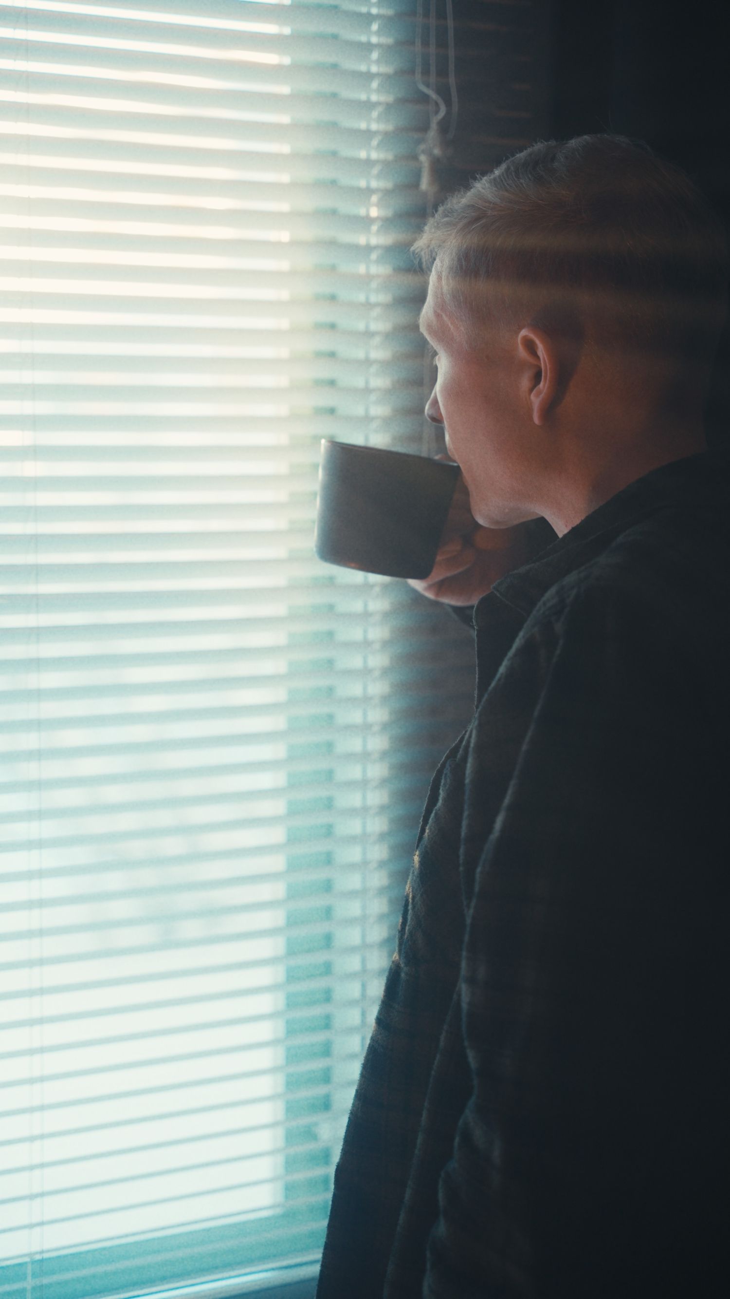 Silhouetted figure drinking from mug while looking through window blinds.