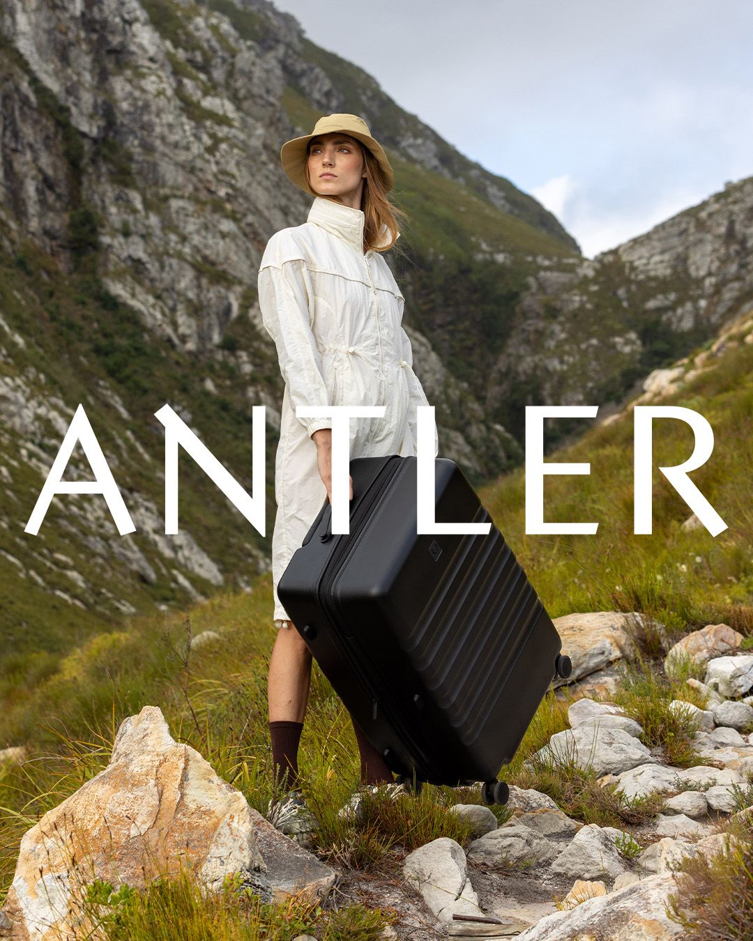 Woman in white dress carrying black luggage through scenic mountain landscape with Antler branding.