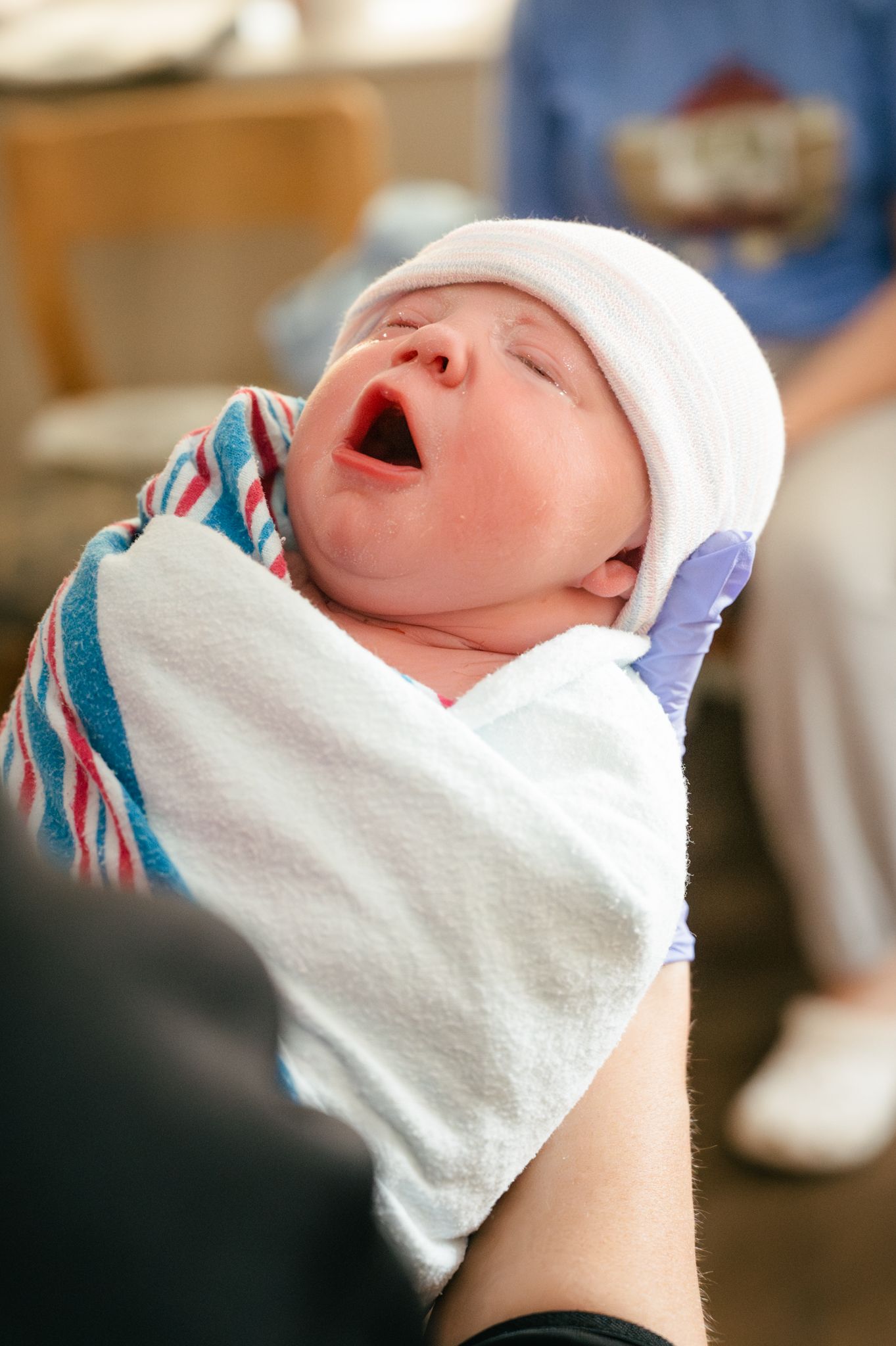 Newborn wrapped in striped blanket and white cap yawns widely.