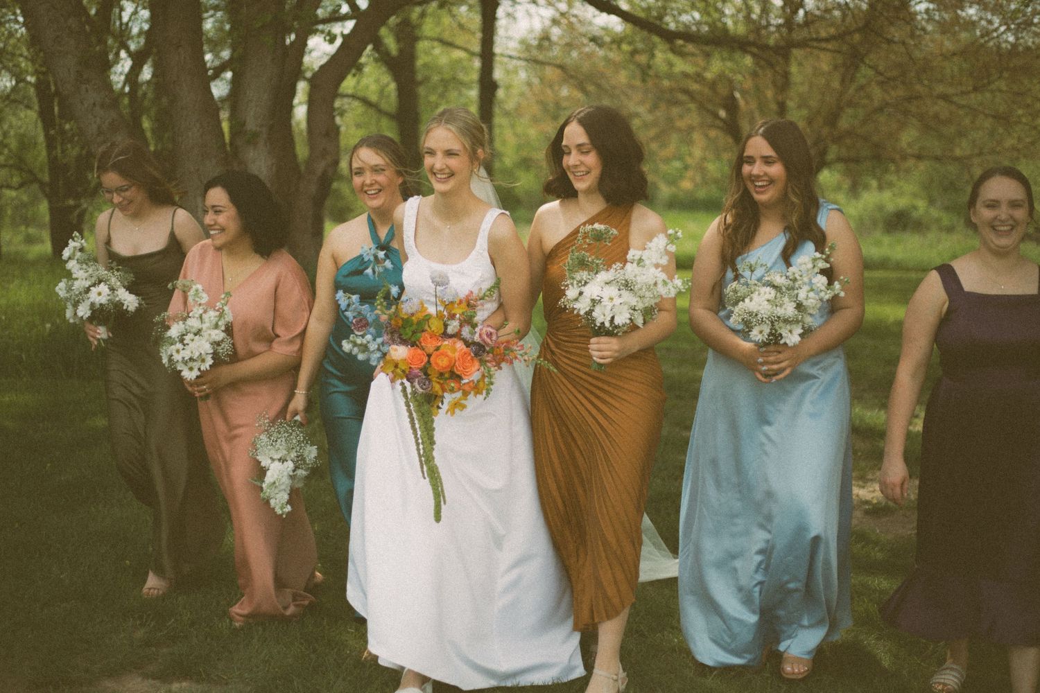 Wedding party with bridesmaids in mixed color dresses walking through wooded area.