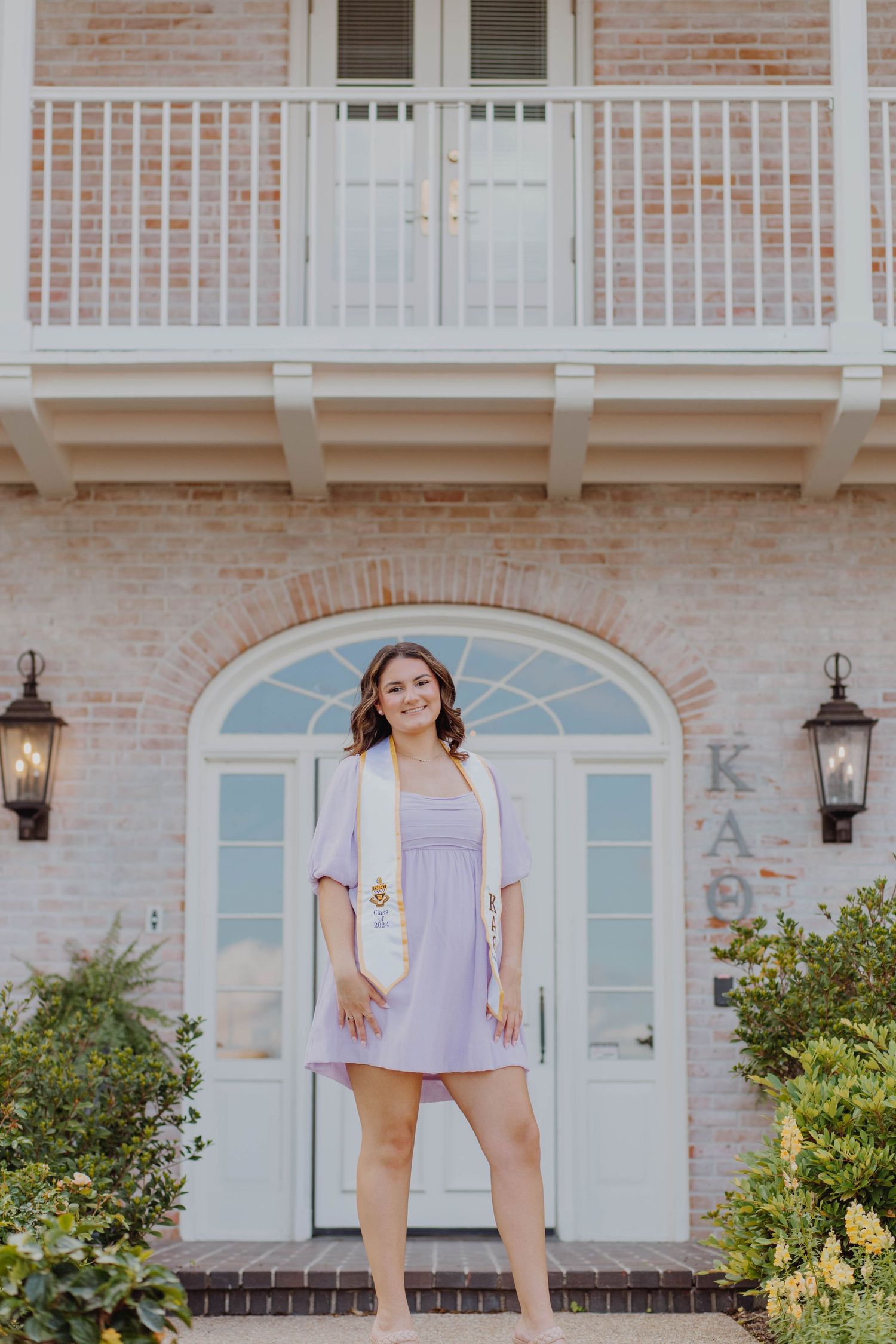 A graduate poses on the steps of a beautiful brick building with white columns and lanterns.