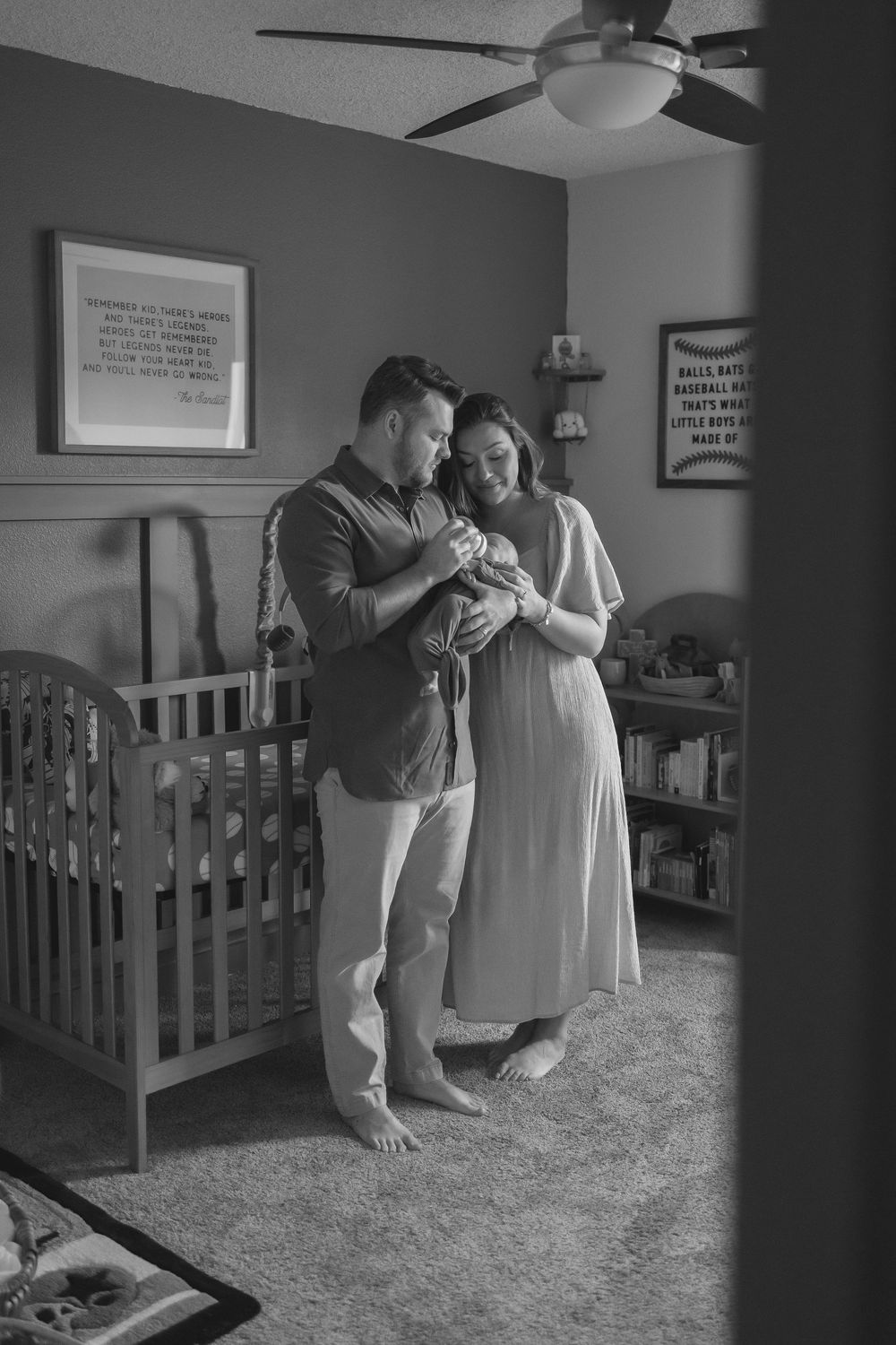 black and white image of a man and woman stood next to a crib in a nursery.  There are stood closely, looking happily at their baby in dads