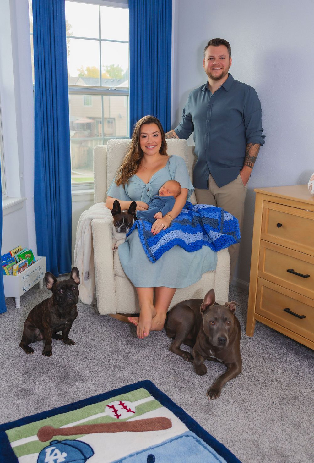 A woman sat on a glider chair in a nursery, holding a baby boy.  Next to her stood up is Dad and 3 dogs
