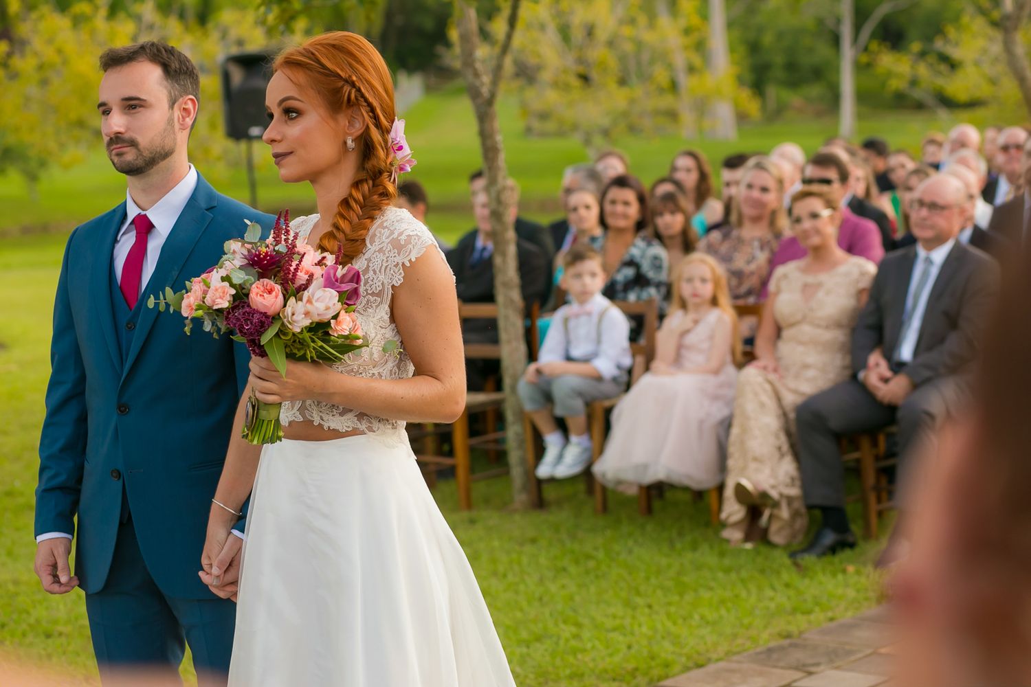 An outdoor wedding ceremony with guests seated on wooden chairs in a garden setting.