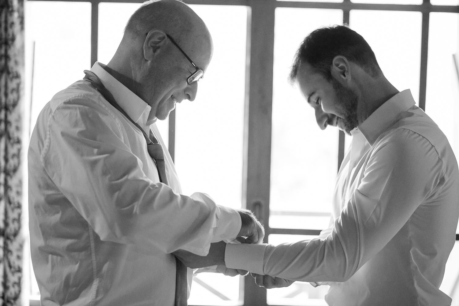 Two people adjusting formal attire while getting ready near a window in black and white.