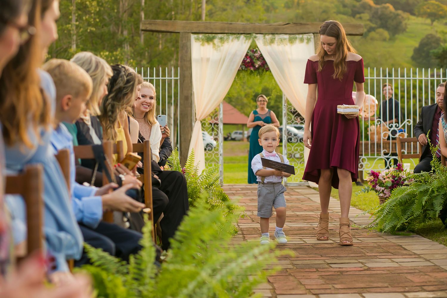 A young ring bearer walks down an outdoor wedding aisle lined with guests and decorative greenery.