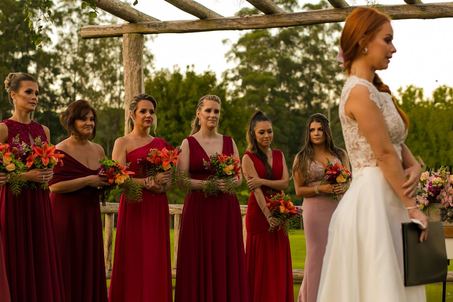 Wedding party stands under wooden pergola with bridesmaids wearing burgundy dresses.