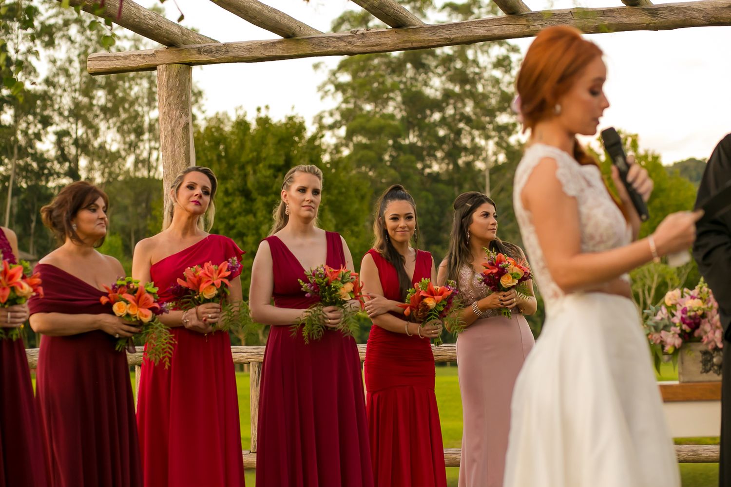 Wedding ceremony with bridesmaids in burgundy dresses holding colorful bouquets under wooden arbor.