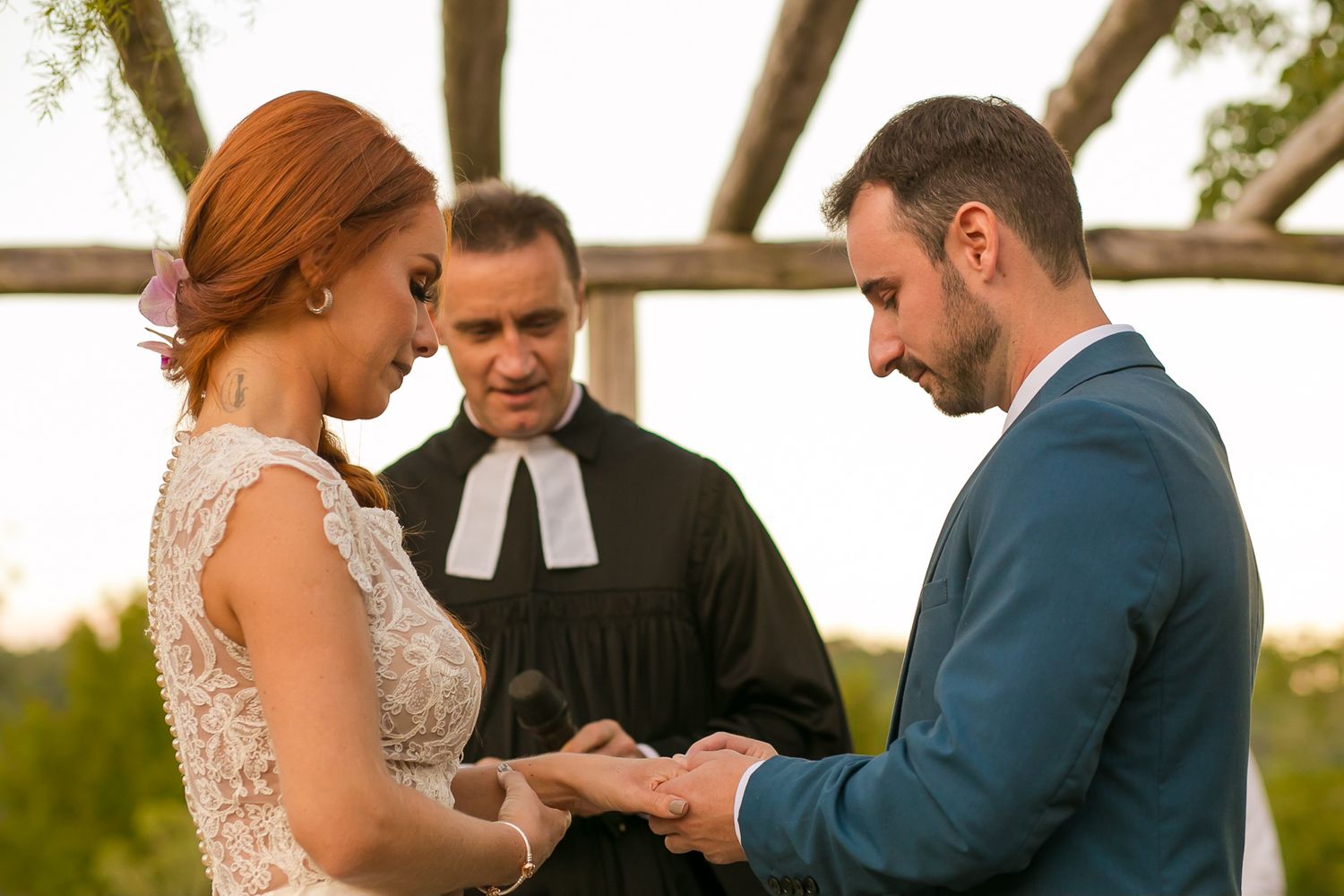 A couple exchanges rings during an outdoor wedding ceremony under a wooden arbor.