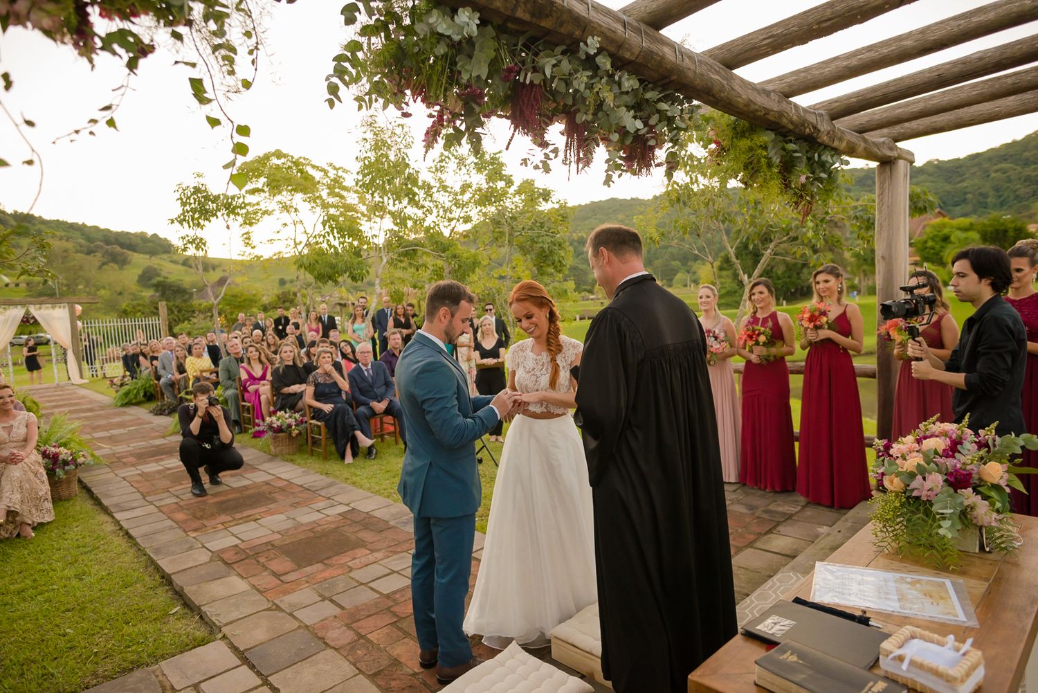 An outdoor wedding ceremony with guests seated on a brick patio surrounded by nature.
