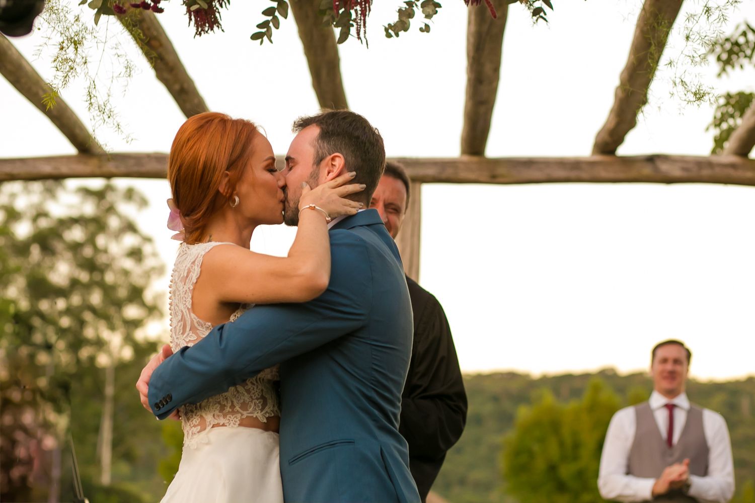 A romantic first kiss at an outdoor wedding ceremony under a wooden pergola.