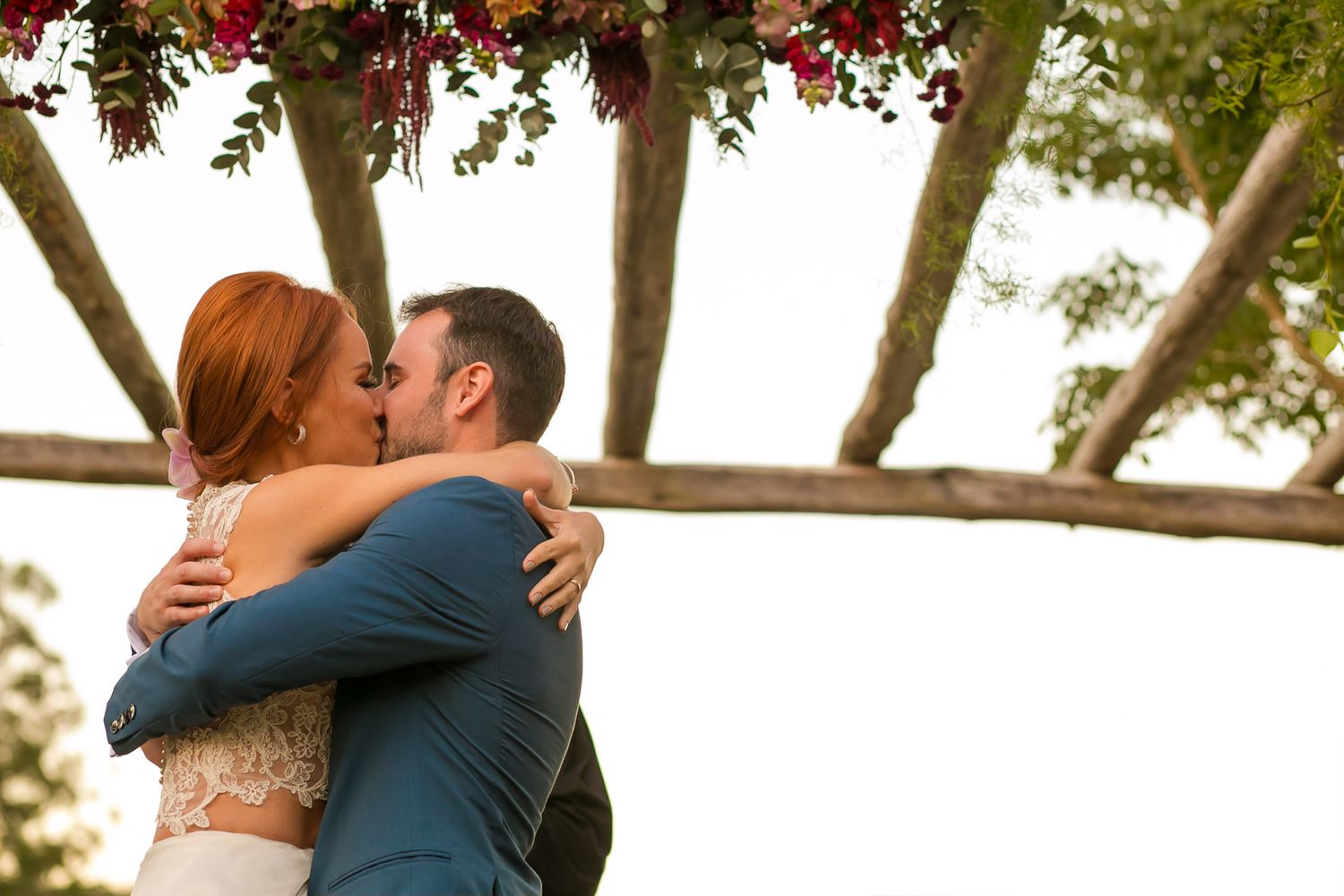 Newlyweds embrace in a passionate kiss under a rustic wooden pergola.