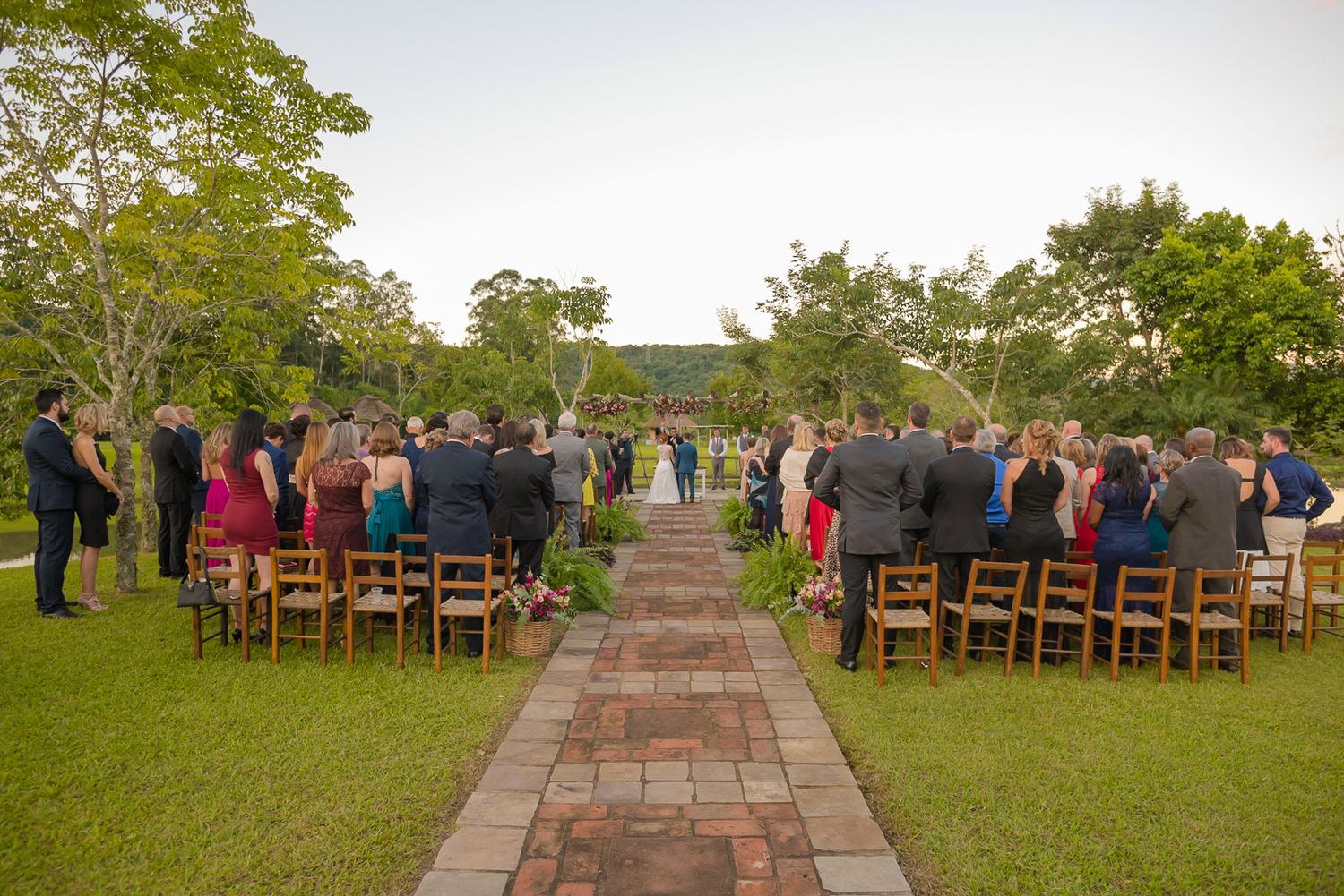 Wedding guests seated in rows on wooden chairs along a brick pathway for an outdoor ceremony.