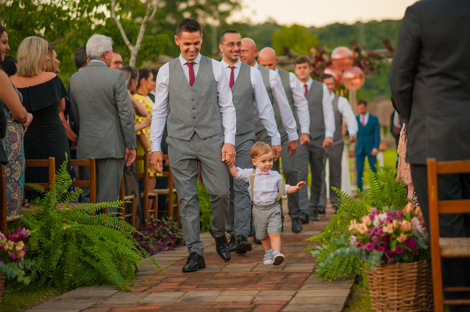 A small child in formal attire walks hand in hand with groomsmen down an outdoor wedding aisle lined with ferns.