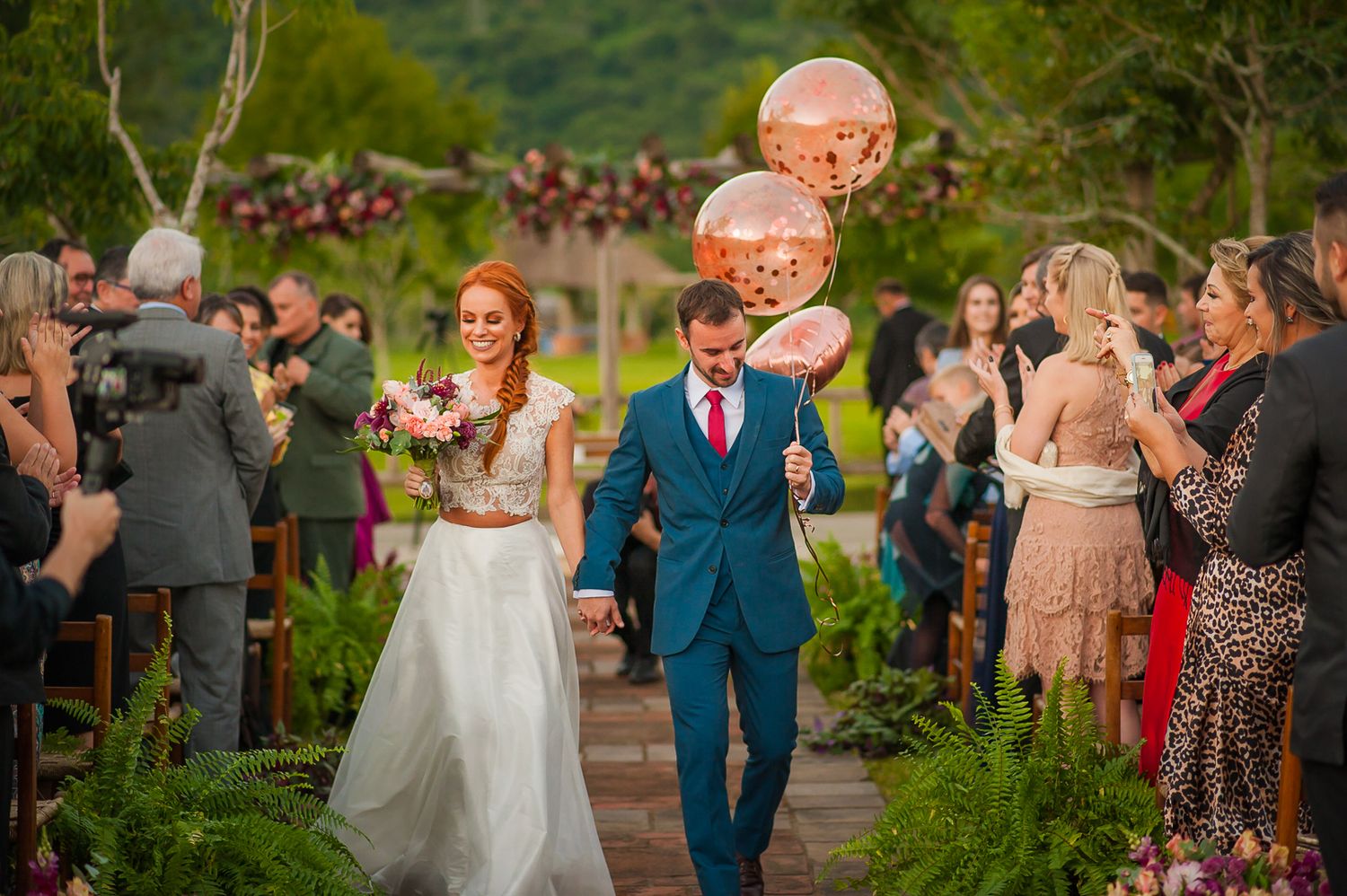 Newlyweds walk down an aisle with pink balloons while wedding guests celebrate.