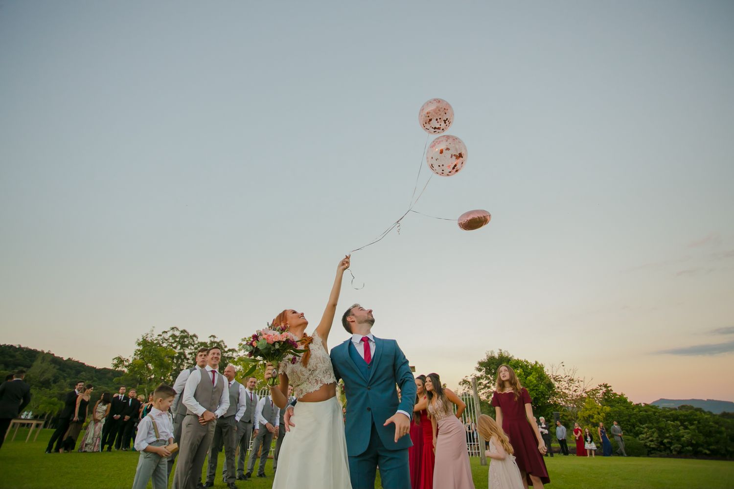 Wedding couple holds pink balloons during sunset celebration with guests.