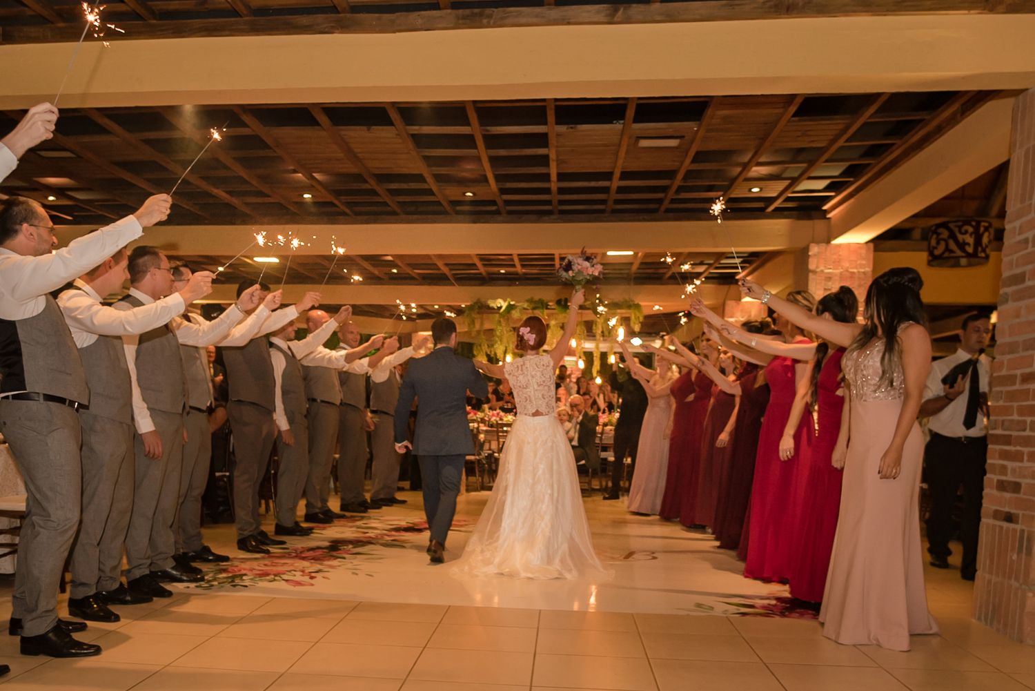 Wedding guests form tunnel with raised arms as couple walks through indoor reception space.
