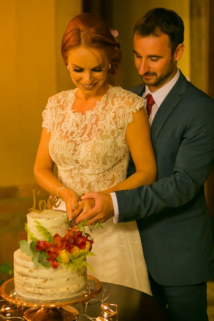 Couple cuts elegant white wedding cake decorated with fresh fruit in warm lighting.
