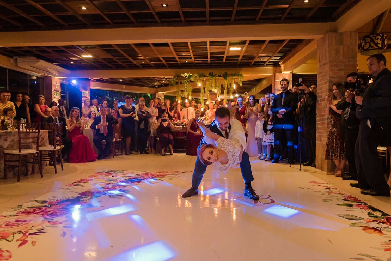 A couple performs a dramatic dance dip on an illuminated reception floor surrounded by guests.