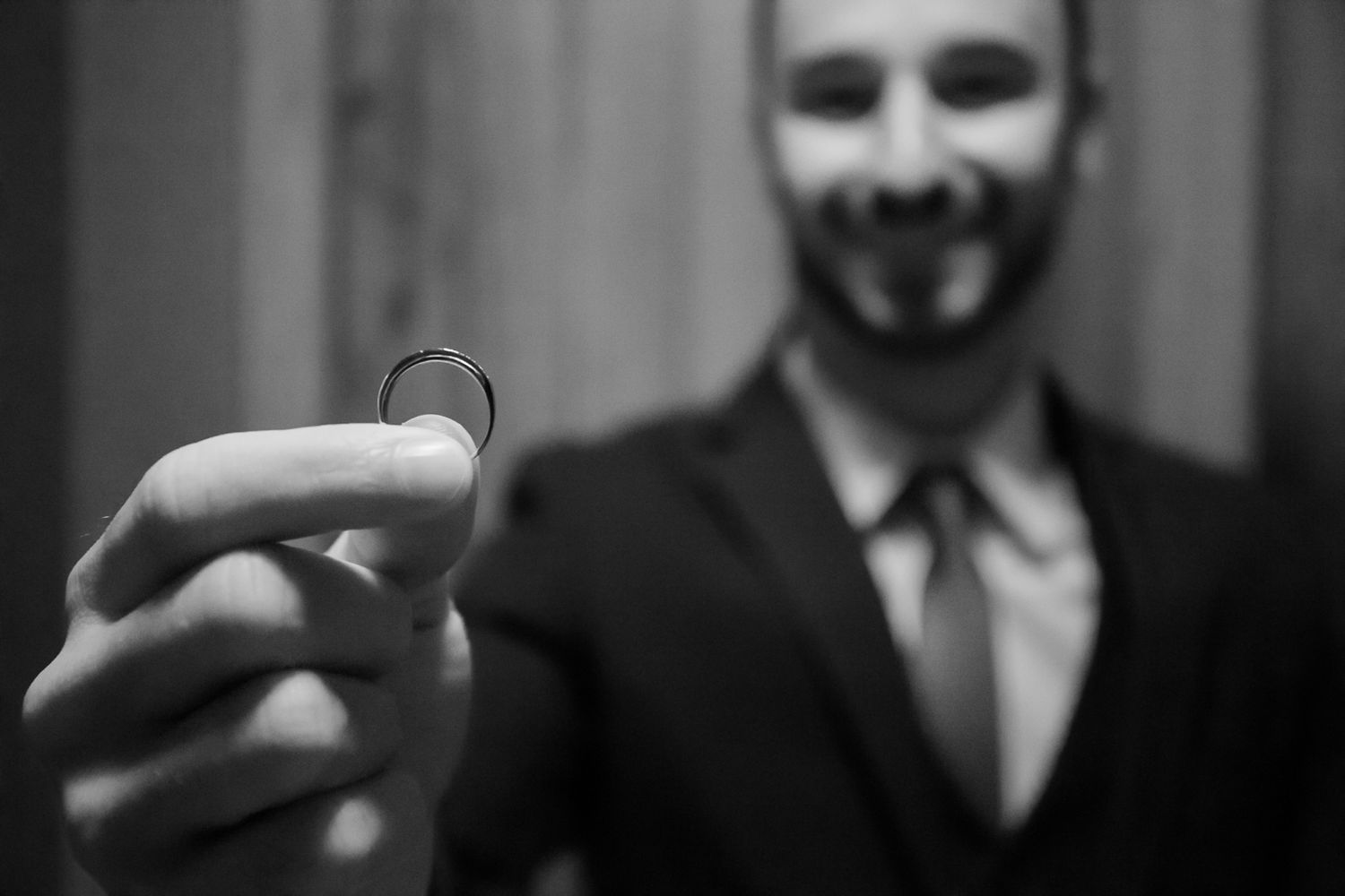 Close-up of a wedding ring being held in focus with suited figure in background.