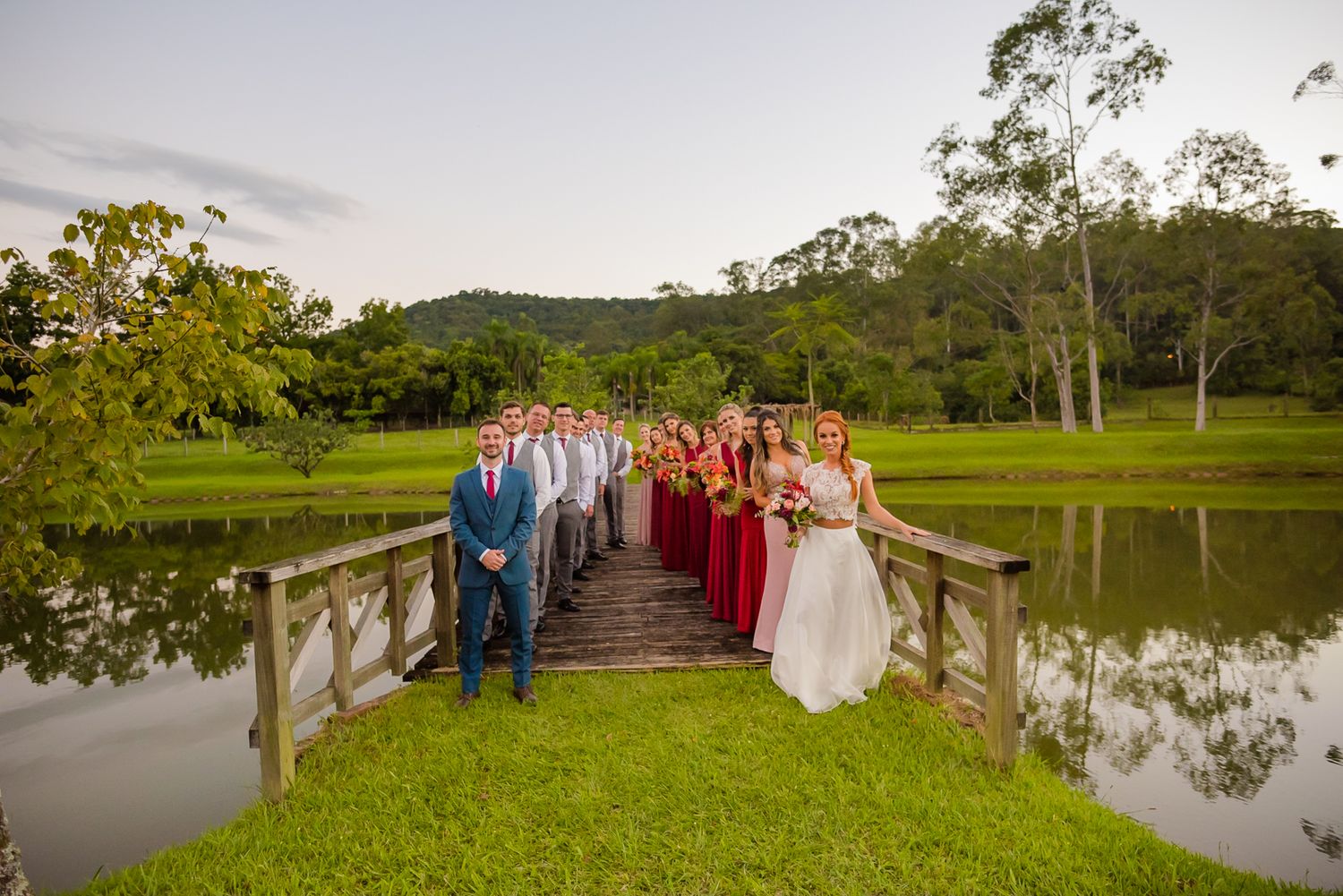 Wedding party gathered on a wooden bridge over tranquil water during sunset.
