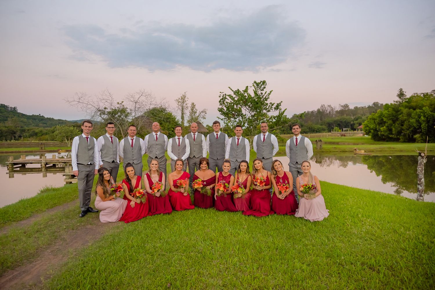 Wedding party in red dresses and gray suits posing together on grassy lawn.