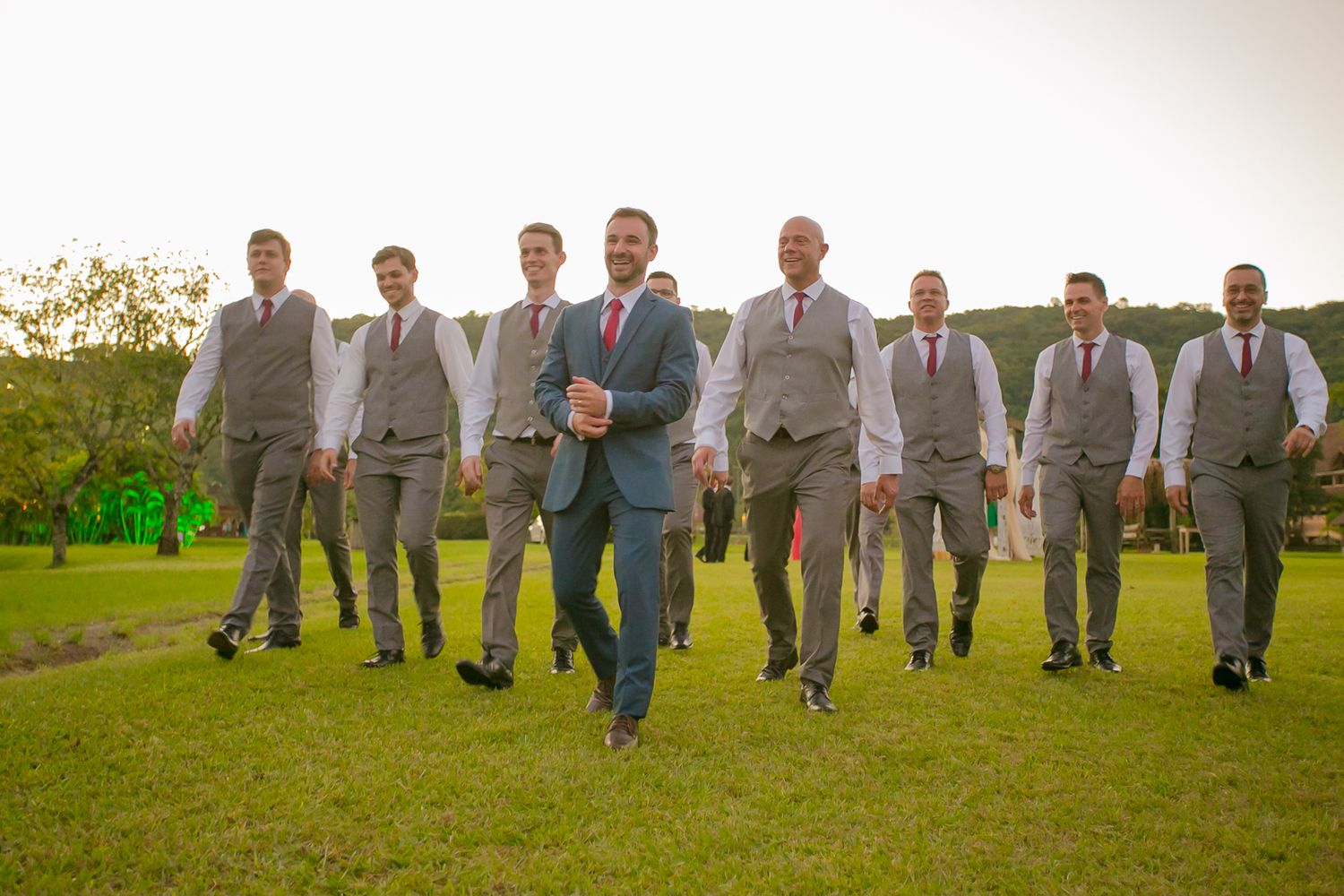 Groomsmen in gray suits walking together across green lawn at outdoor wedding.