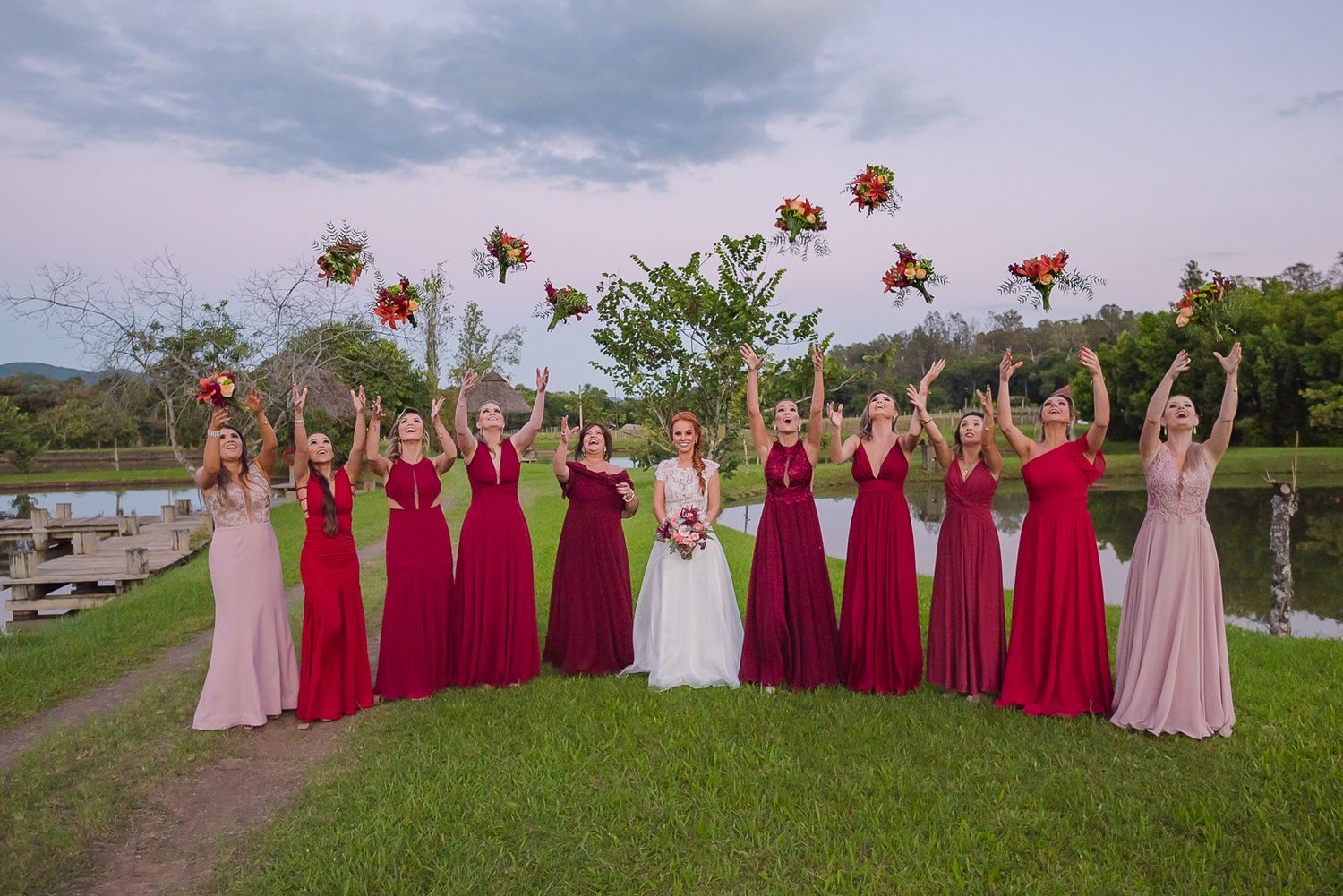 Bridesmaids in red dresses tossing bouquets in the air against cloudy sky.