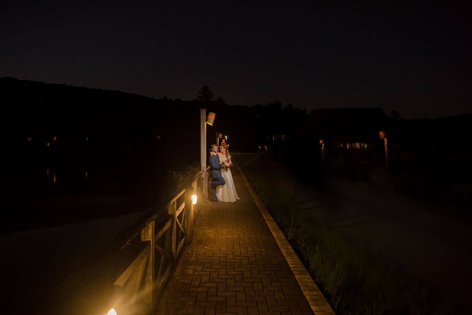 A dimly lit wooden bridge pathway illuminated by lanterns at night.