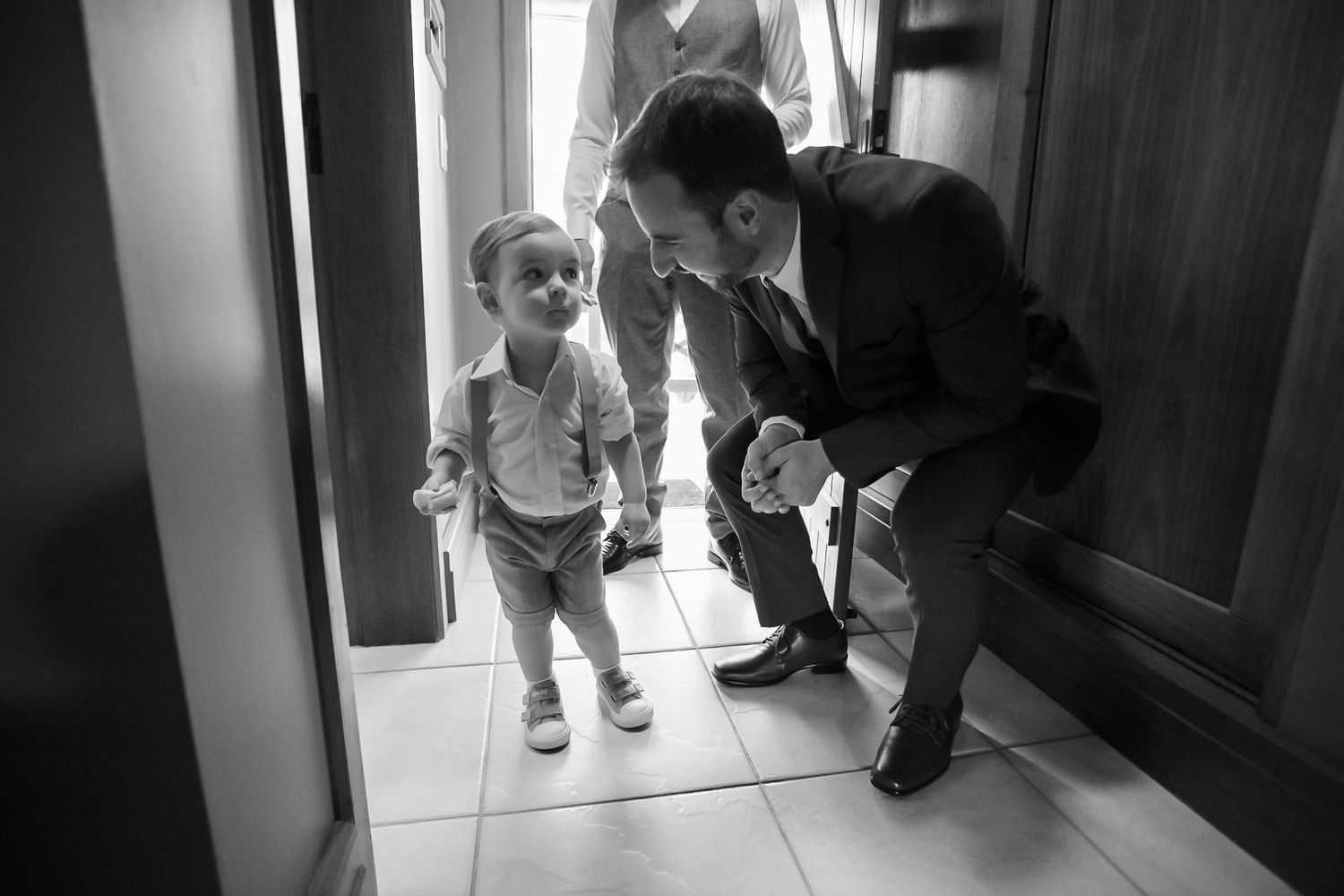 A candid black and white photo shows a toddler entering a doorway while adults in formal attire look on.