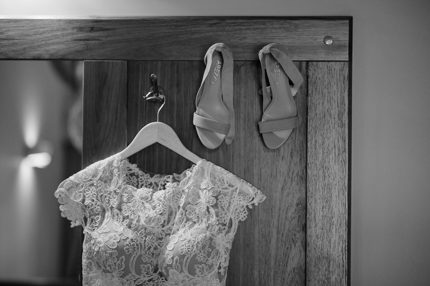 A delicate lace wedding dress and sandals hang on wooden hangers against a wall.