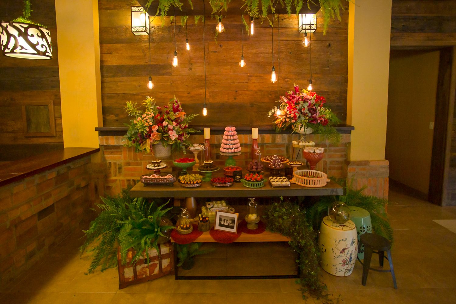 A rustic dessert table display with hanging lights, floral arrangements, and multiple cakes and treats on wooden shelves.