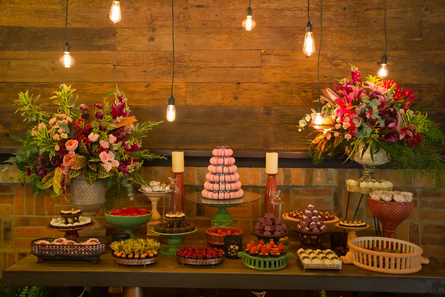 Wedding dessert table with multiple cakes and treats arranged under hanging lights with floral decorations.