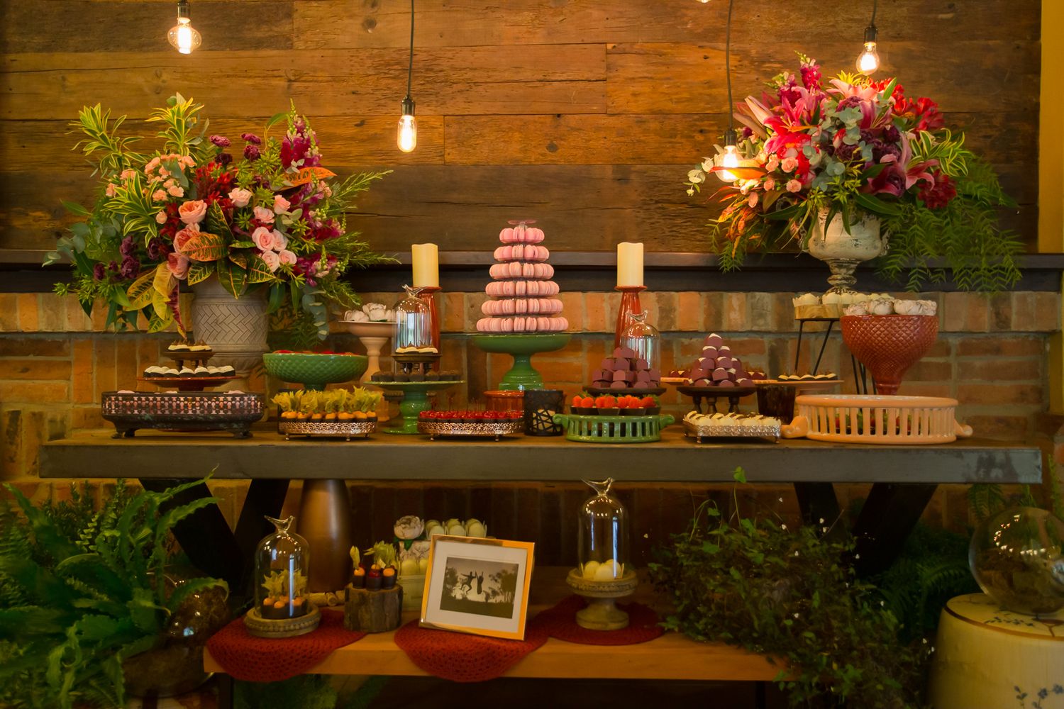 Rustic wedding dessert bar featuring various cakes and treats displayed with string lights and pink floral arrangements.